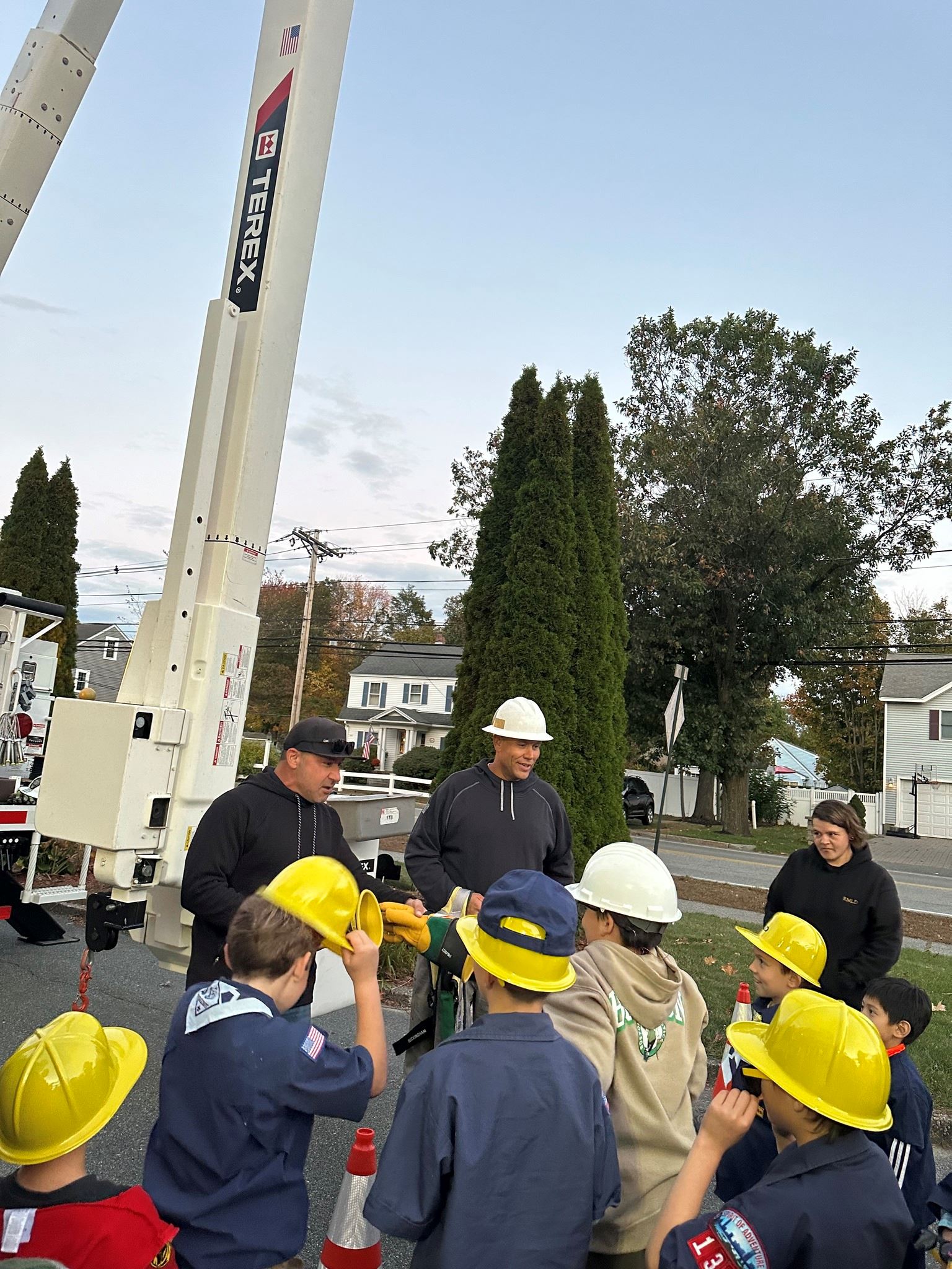 Two lineworkers explain safety to a group of cub scouts wearing yellow hard hats.