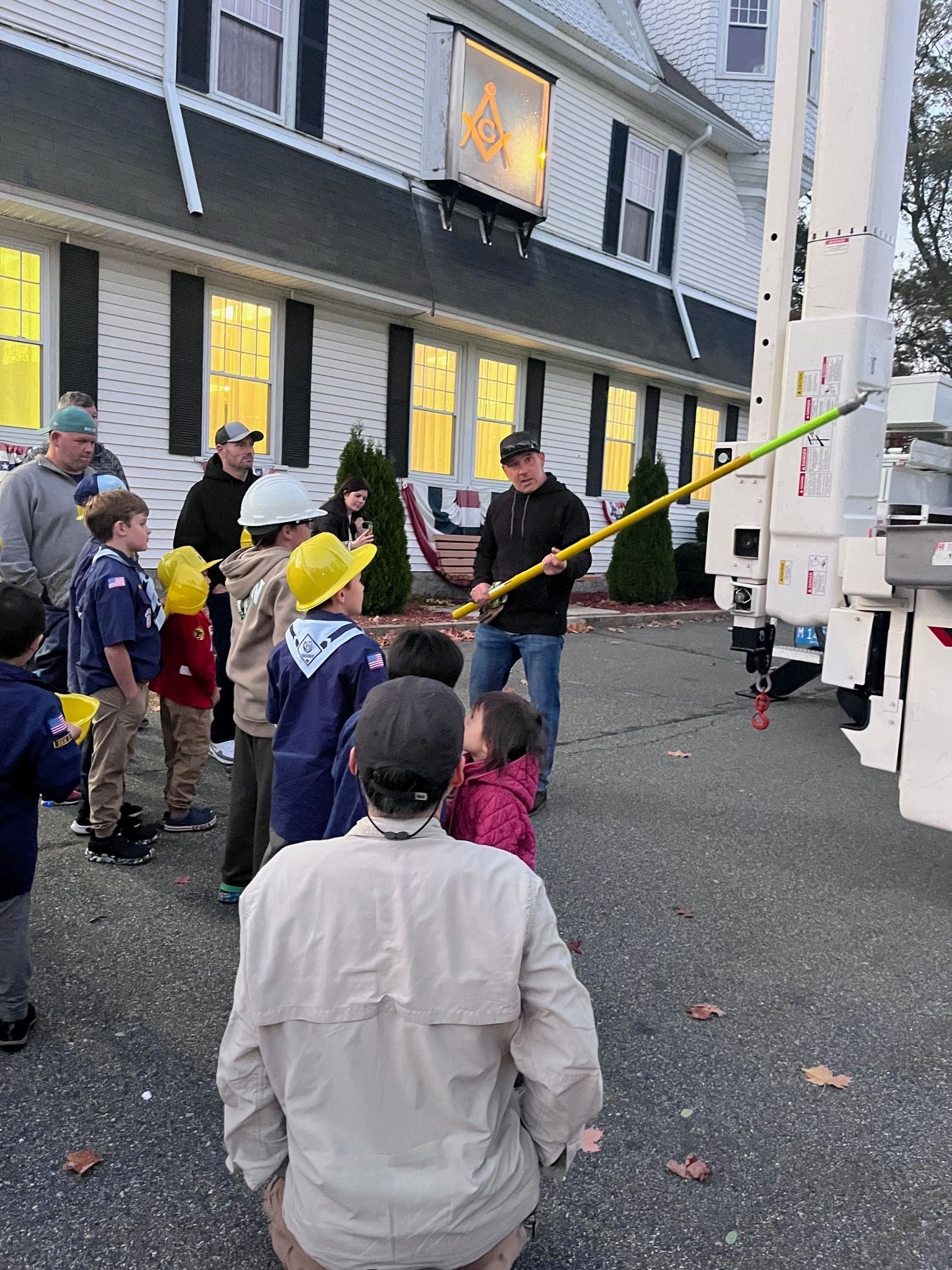 A lineworker explains a hot stick to a group of cub scouts wearing hard hats.